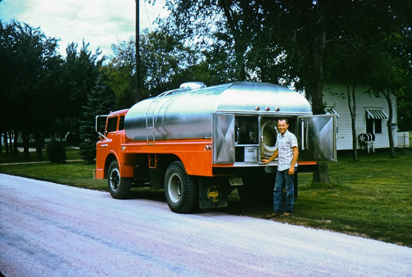 Man in blue jeans and a short sleeve button down shirt stands in the grass next to a residential street. He stands behind an orange truck with a silver tank on the back used to haul milk in the 1960s and 70s parked mostly in the grass at the side of the road. There are open doors on the back of the truck.