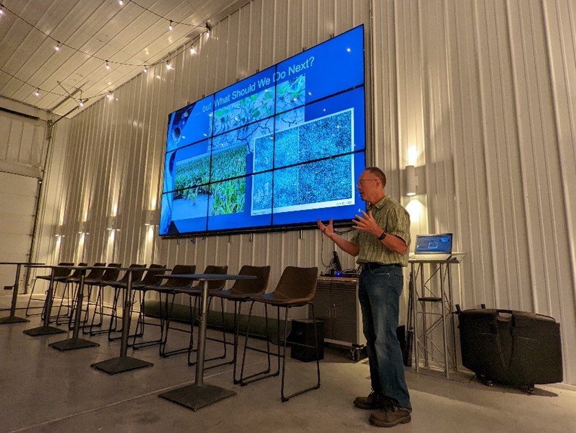 Brad Doorn gestures while presenting satellite imagery on a large digital display wall in a room with high-bar seating.