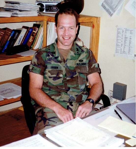 White man sitting at a desk in U.S. Army camouflage uniform. The desk has a pen and various stacks of paperwork and there are shelves behind him with various books and a stereo.