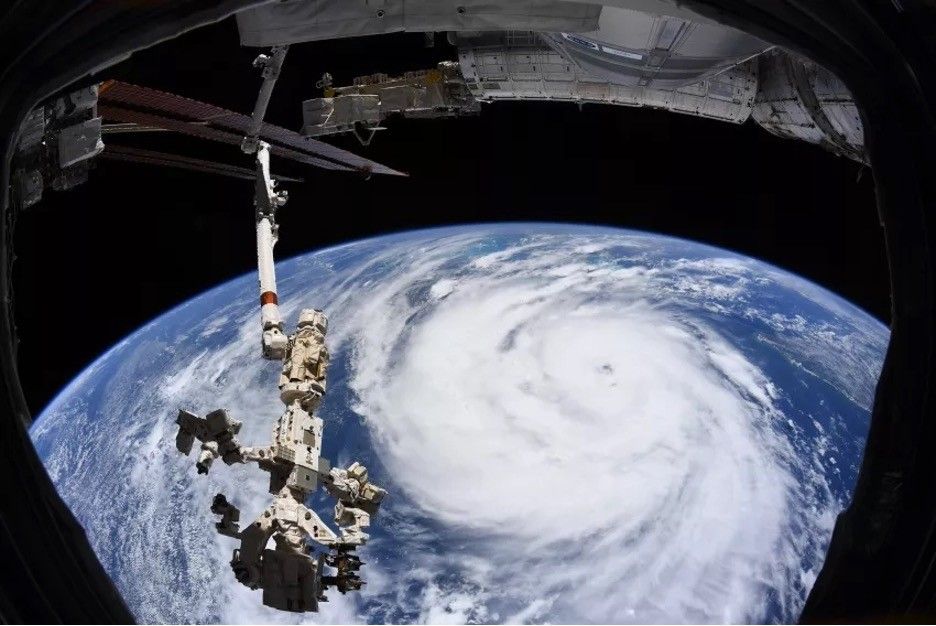 An image of Earth from space showing the swirling clouds of a hurricane forming over the ocean with a robotic arm extending from the International Space Station.