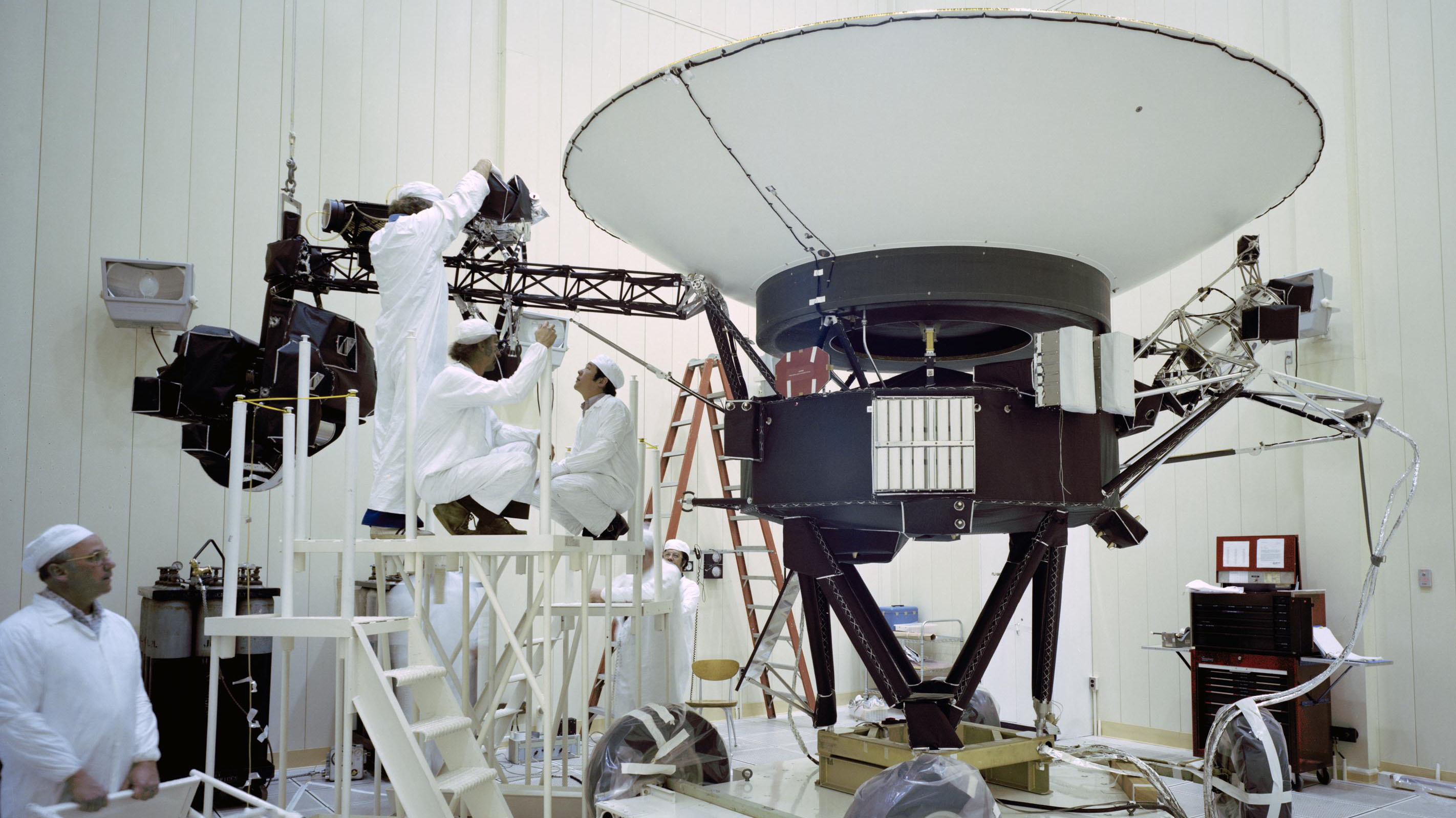 Three workers dressed in white suits in a clean room are on a scaffolding mid-frame, tending to a large, black metal spacecraft. Its large, white radio dish is above them, extending upward toward the ceiling. A fourth worker, similarly dressed, stands at left, on the floor, watching.