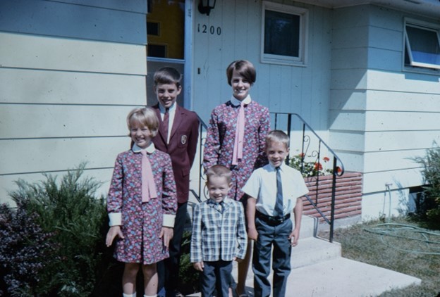 Five children—young Brad Doorn and four siblings—stand outside a house. Two girls wear matching floral dresses, and the three boys are in various formal attire.