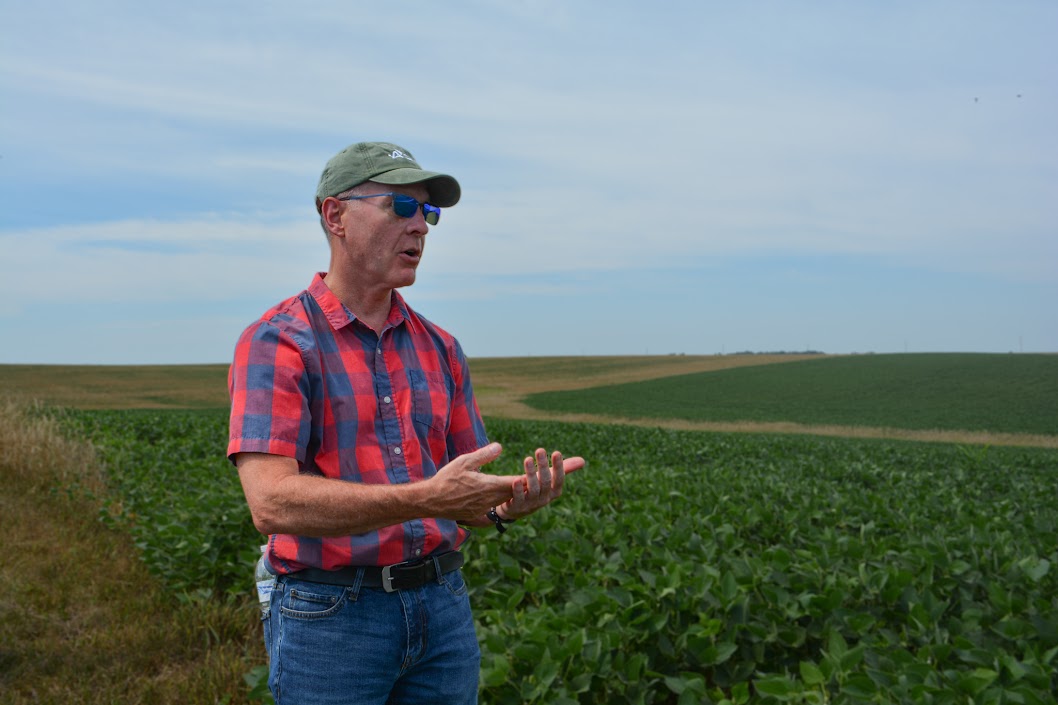 A man stands in a crop field, wearing a green cap, sunglasses, and a red and blue plaid shirt. He is mid-sentence, gesturing with his hands, against a backdrop of green farmland under a partly cloudy sky.