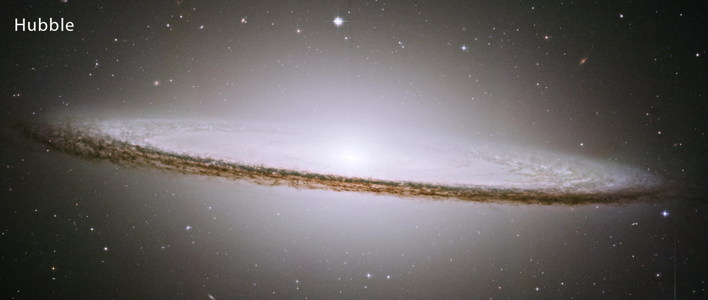 Hubble’s view of the Sombrero galaxy. In this view, the galaxy is an oblong, pale white disk with a glowing core over the inner disk. The outer disk is darker and clumpy.