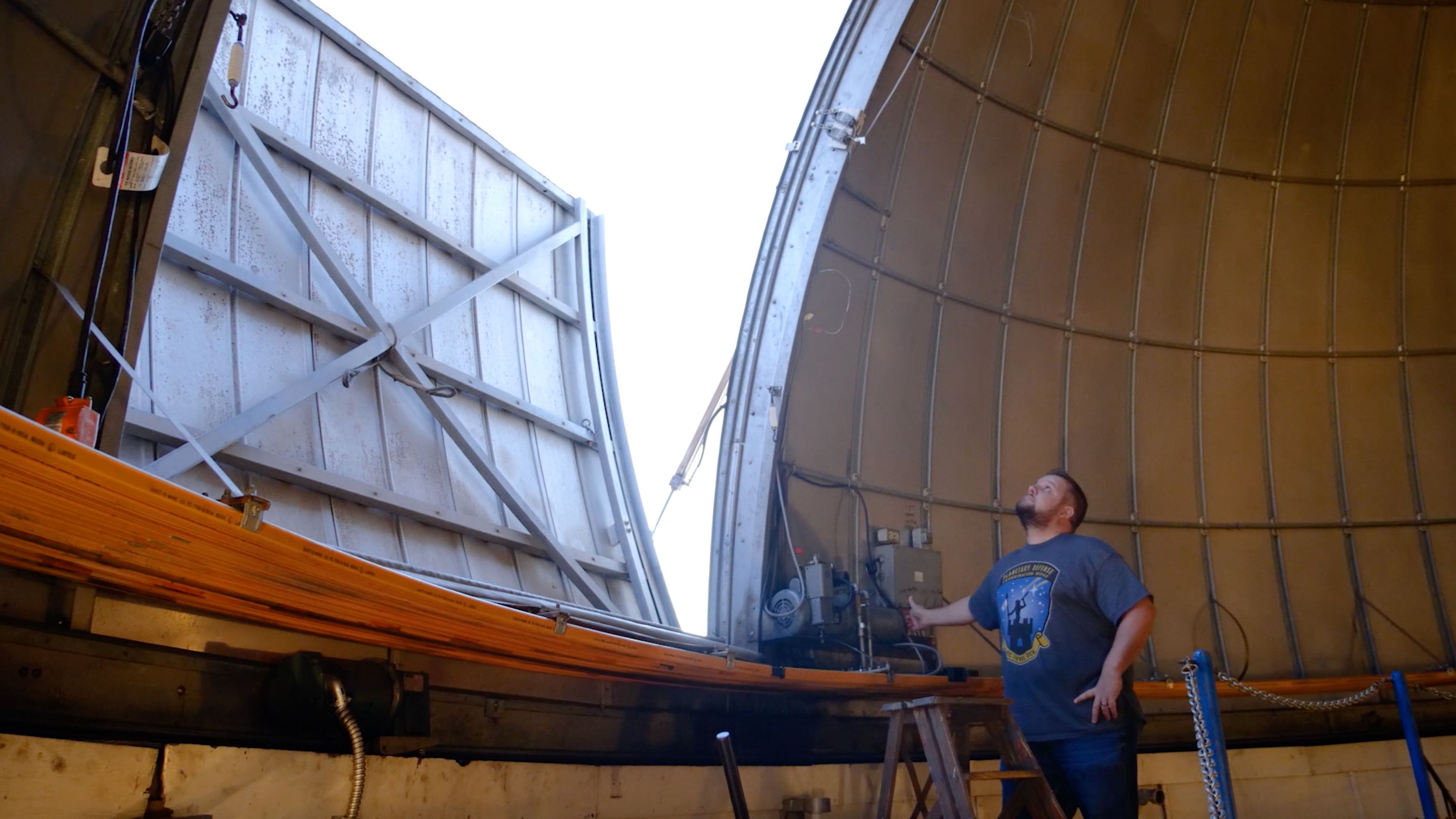 The image depicts the interior of an observatory dome with a large opening through which daylight is visible. The dome structure consists of curved metallic panels that meet at the top, with one panel slightly open on the left side, revealing a cross-braced metal framework. In the foreground, a person stands on a ladder inside the dome, wearing a casual T-shirt with a prominent logo. The person is looking upward toward the opening, partially illuminated by the ambient light. Behind them is an array of mechanical equipment, possibly part of the dome’s operation system. The overall color scheme is composed of metallic grays and earth tones from the dome's surfaces.