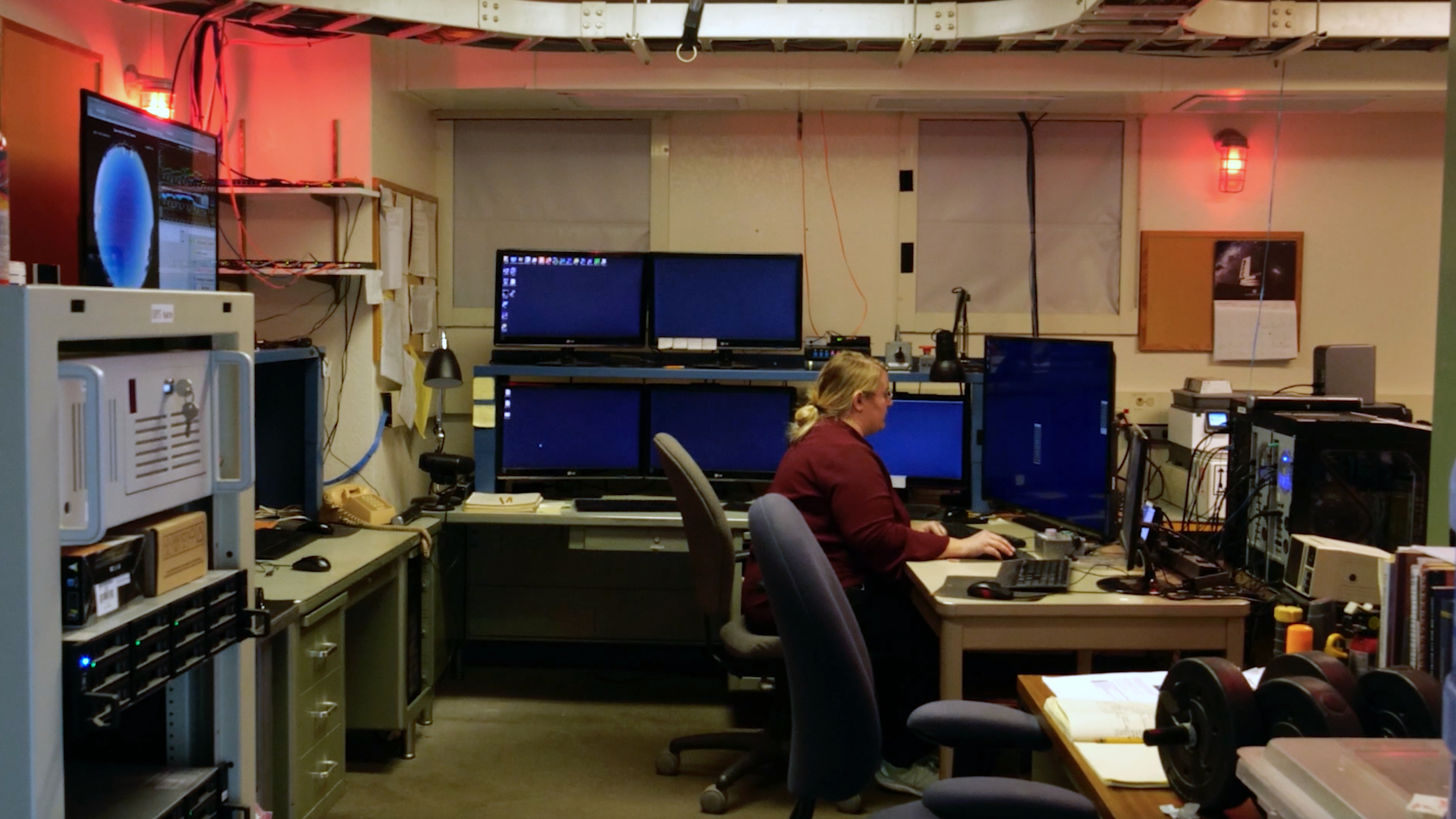 The image depicts a dimly lit control room with a red overhead light casting a subtle glow. On the right, a person with light hair is seated at a desk with six computer monitors, some displaying a blank dark screen, while others have windows open but with indistinguishable content. The person is focused on the central monitors and using a keyboard. On the left, a larger console with a visible blue screen displaying a circular graphic is mounted slightly elevated. The room is filled with various electronic equipment, shelves with stacked papers, and wires hanging from the ceiling. A lamp is positioned on the desk, providing additional light. In the background, a corkboard with a calendar and a small poster hangs on the beige wall.