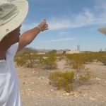 The image shows a man in the foreground wearing a wide-brimmed, light beige hat and a white polo shirt. He is pointing towards the distance with his right arm extended. The landscape is a desert environment with sparse vegetation and rocky ground. In the background, there is a large radio antenna or satellite dish, characterized by its wide, circular dish supported by a strong base. The sky is clear, with a few wispy clouds scattered across it. The horizon shows low hills or mountains in the distance.