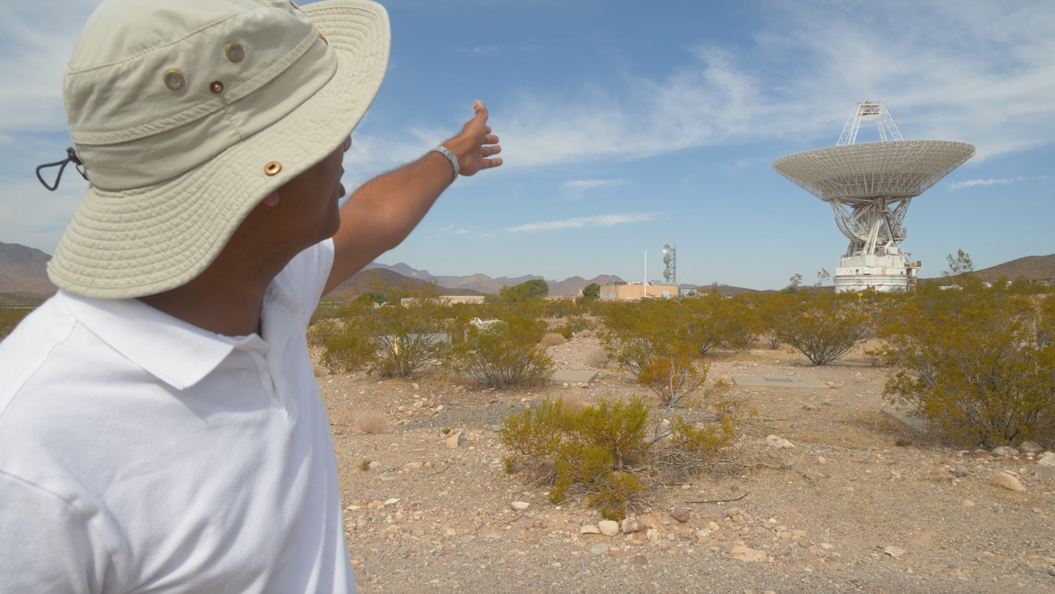 The image shows a man in the foreground wearing a wide-brimmed, light beige hat and a white polo shirt. He is pointing towards the distance with his right arm extended. The landscape is a desert environment with sparse vegetation and rocky ground. In the background, there is a large radio antenna or satellite dish, characterized by its wide, circular dish supported by a strong base. The sky is clear, with a few wispy clouds scattered across it. The horizon shows low hills or mountains in the distance.