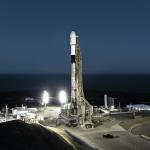 A SpaceX Falcon 9 rocket sits on a launch pad at dawn. Lights illuminate the rocket from below and the sky is just beginning to lighten on the horizon.
