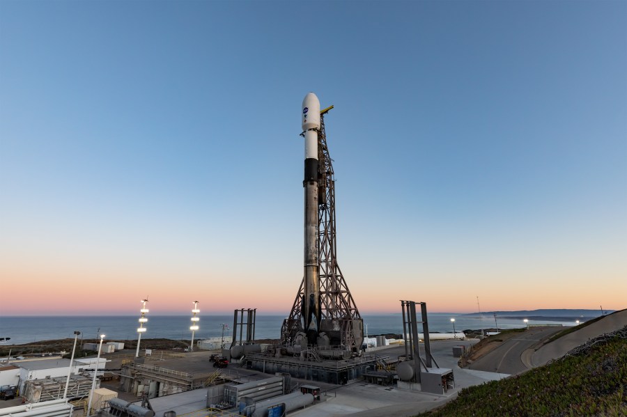 A SpaceX Falcon 9 rocket stands erect on the launch pad along side a retractable gantry. The horizon is golden in the light of dawn with a deeper blue sky above.