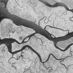 A black-and-white synthetic aperture radar (SAR) satellite image captured in landscape orientation showing a frozen river system. A large, bright ice- and snow-covered landmass dominates the center and right of the frame, its edges defined by sharp contrast against dark surrounding water. Intricate drainage channels and surface texture are visible across the ice surface. Dark water areas in the lower left display swirling, layered patterns indicative of slow-moving or partially frozen flow. A small airstrip is visible on an island in the left portion of the frame.
