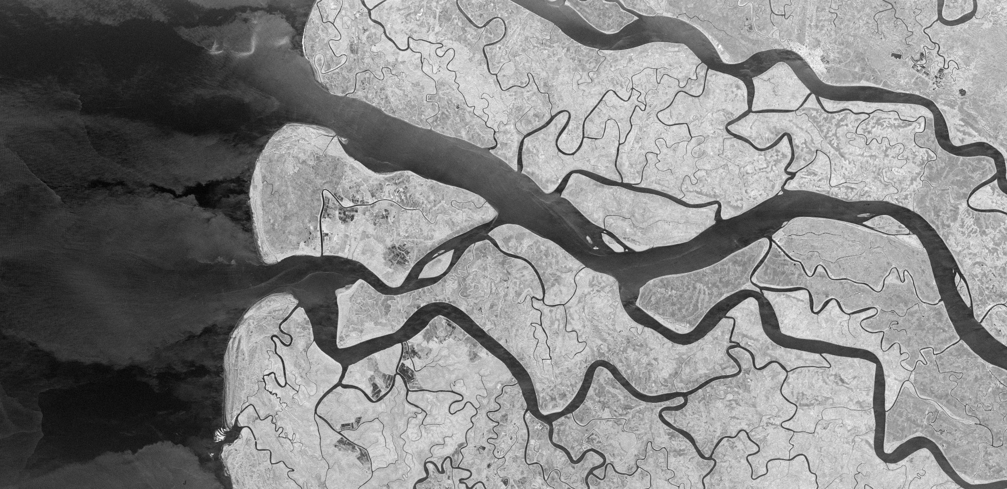 A black-and-white synthetic aperture radar (SAR) satellite image captured in landscape orientation showing a frozen river system. A large, bright ice- and snow-covered landmass dominates the center and right of the frame, its edges defined by sharp contrast against dark surrounding water. Intricate drainage channels and surface texture are visible across the ice surface. Dark water areas in the lower left display swirling, layered patterns indicative of slow-moving or partially frozen flow. A small airstrip is visible on an island in the left portion of the frame.