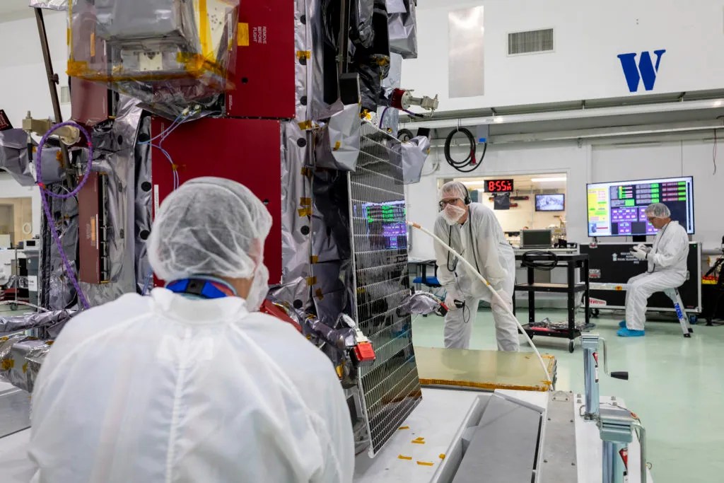 Technicians in cleanroom suits work on a spacecraft inside a high-tech laboratory. One technician holds a tool near a solar panel on the spacecraft while another monitors data on a computer screen. The spacecraft is partially wrapped in protective foil and wiring. A digital clock on the wall reads 8:55.
