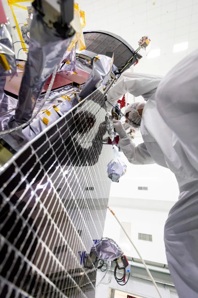 Close-up, low-angle view of a technician in a cleanroom suit working on the side of a spacecraft. The technician is inspecting or securing components near a solar panel, which dominates the foreground of the image. The spacecraft is covered in reflective insulation and intricate wiring. The bright, sterile environment of the cleanroom is visible in the background.
