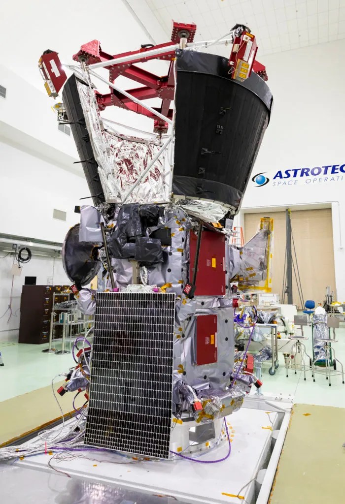 A spacecraft in a cleanroom at Astrotech Space Operations is shown undergoing final preparations. The spacecraft is wrapped in thermal insulation and outfitted with red and silver components, wiring, and a large black sunshield structure at the top. A solar panel is mounted on the lower half, and various instruments and structural supports are visible around the spacecraft.
