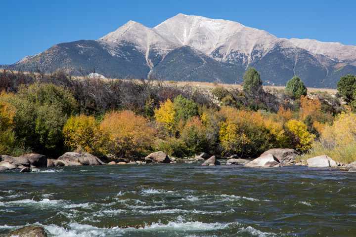 A ground-based photo shows the range of landscapes within the national monument. It includes rough river water in the foreground, shrubby vegetation in the middle, and mountainous terrain in the background.