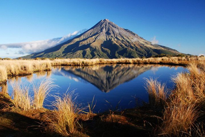 Photograph of Mount Taranaki taken from the side showing the snow-free flanks and peak. The mountain is reflected in the waters of a small lake that is encircled by light brown grasses.