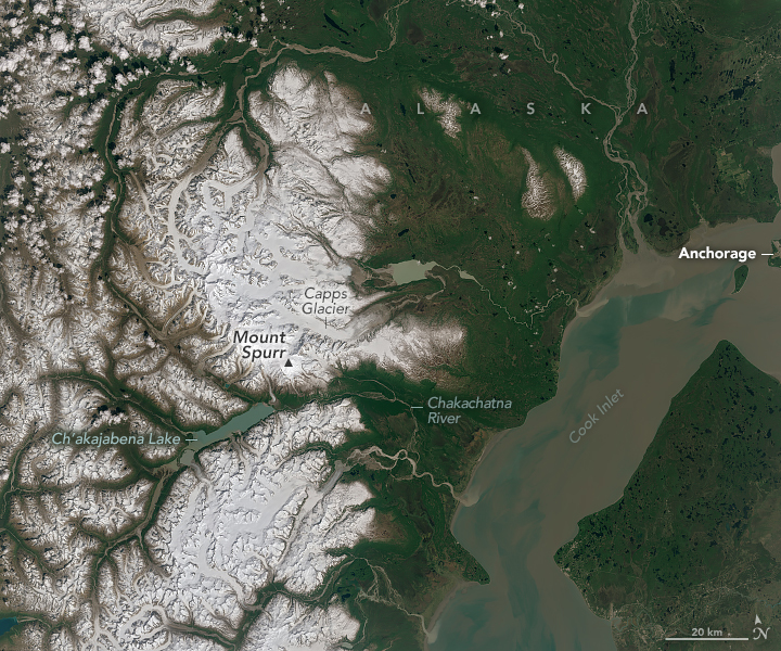 A wider view of the same location shows Mount Spurr toward the middle-left and the edge of Anchorage on the right. The left part of the image is mountainous and white, whereas the right side is primarily green. The blue-brown waters of Cook Inlet cut through the bottom right.