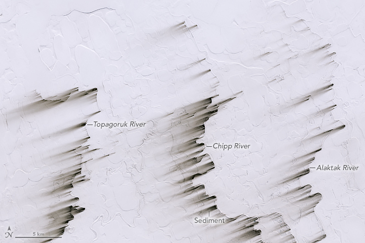 A satellite image shows snow-covered land with the shapes of lakebeds and riverbeds faintly visible. Parallel dark streaks extend toward the left-bottom left of the image from some parts of three riverbeds.
