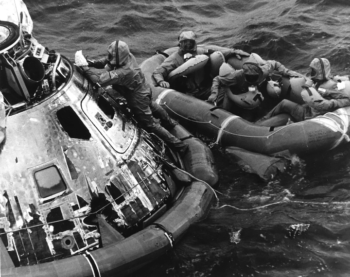 A black-and-white photo taken in 1969 shows the astronauts waiting in a raft for helicopter pickup after exiting the Columbia command module. They are wearing hazmat-like suits called biological isolation garments (BIGs) to prevent the spread of potential lunar pathogens.