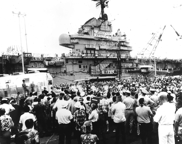 A black-and-white photo taken in 1969 shows the USS Hornet at a pier in Pearl Harbor. A large crowd has gathered around the aircraft carrier to welcome the Apollo 11 astronauts.