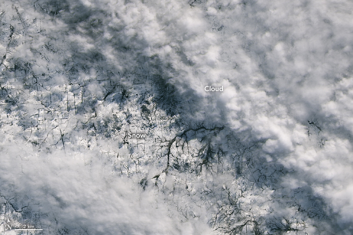 A satellite image shows a more detailed view of fresh snowfall on the Northern Tablelands region of New South Wales, Australia. Bands of white puffy clouds extend across the upper right and lower left. In the middle of the image, white snow-covered ground is broken up by several rivers and dark lines separating fields.