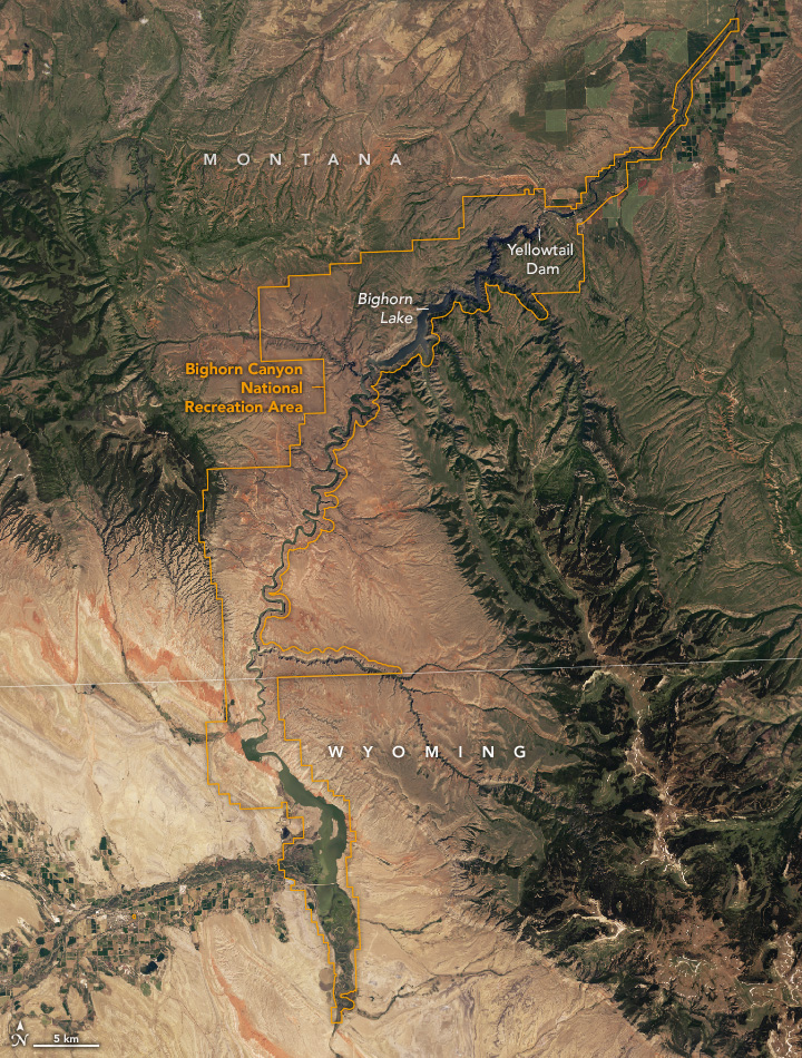 The outlines of Bighorn Canyon National Recreation Area are shown on a satellite image of northern Wyoming and southern Montana. The winding canyon runs roughly north-south through the scene. Some of the land on either side is reddish-tan and some is green and mountainous. A sharply folded reddish rock layer is visible to the left of the recreation area.