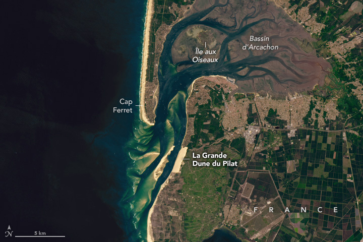 A satellite image shows an area of coastal France, where a sandy beach is interrupted by a channel leading to a tidal bay. This estuary contains an island and marsh areas with branching channels of darker water. South of the bay along the coast is a bright tan block, which is a large sand dune. Areas of forest and rectangular agricultural fields are inland.