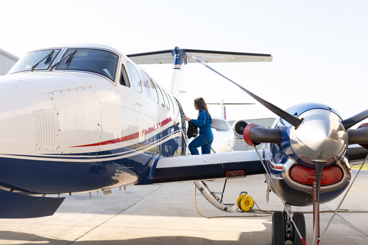 A small aircraft sits on the tarmac and is shown from the front. A woman in a blue flight suit is boarding the plane.