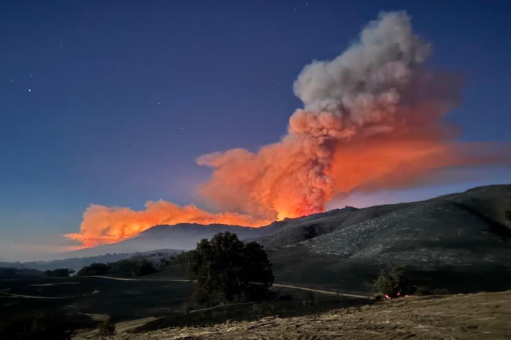 A photograph taken from the ground features a towering cloud of smoke rising from behind a ridge. Much of the smoke plume glows orange in a sign of the active fire burning out of view beneath the ridgeline.