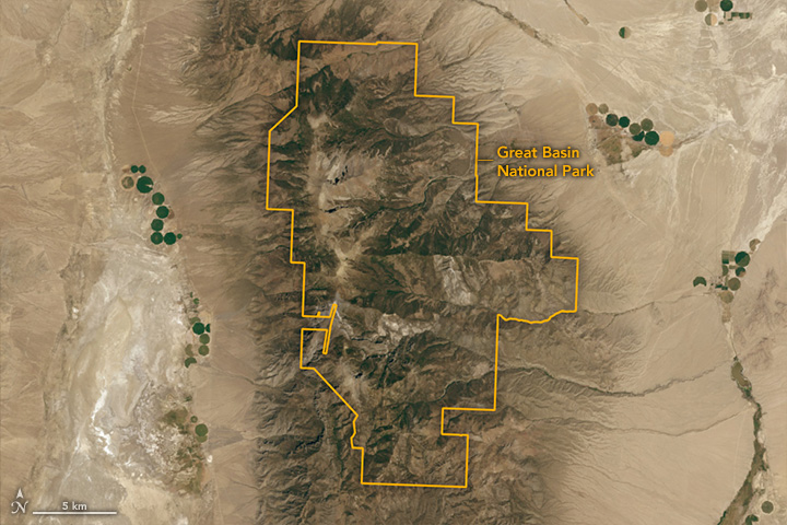 A satellite image shows Great Basin National Park in the daytime. The right and left thirds of the image show a tan-colored desert landscape containing some round, green agricultural fields. The middle part, which contains the park, is mountainous.