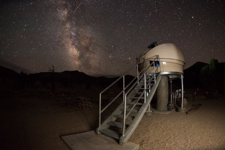 A ground-based photo taken at nighttime shows a dome-shaped astronomical observatory in the foreground with the silhouette of mountainous terrain in the background. The night sky shows an abundance of stars, the streak of a meteor, and the band of light from the Milky Way.