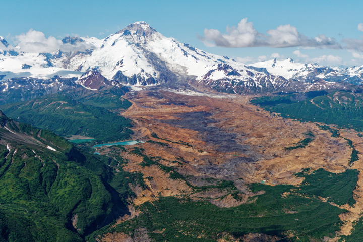 The snow-covered Iliamna Volcano appears in the distance in this photograph, and the Red Glacier flows into a valley in the foreground. One slope of the mountain is covered in rocky debris. The valley is mostly free of vegetation and is full of hummocky piles of tan, reddish, and dark brown rock or soil.