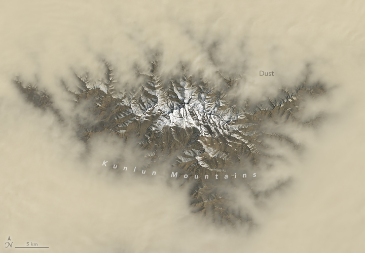 A more detailed view reveals a blanket of dust with a gap at its center, through which snow-capped mountains are visible. Textured patterns in the dust layer appear to be shaped by the underlying terrain.