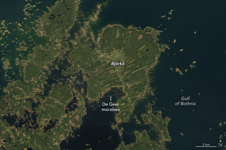A detailed satellite image of the island of Björkö in Finland's Kvarken Archipelago. The island appears to be divided into elongated fields colored tan and different shades of green. Glacial features called De Geer moraines are visible around the coastline as narrow parallel islands.