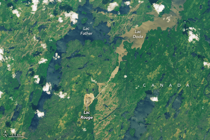 A small, roughly oval-shaped lakebed is in the center of this satellite image of Quebec. A tan channel runs northeast from the lakebed until it meets a large lake that runs along the left and top sides of the image. To the left of the channel, the lake water appears dark blue, and to the right, it appears tan.