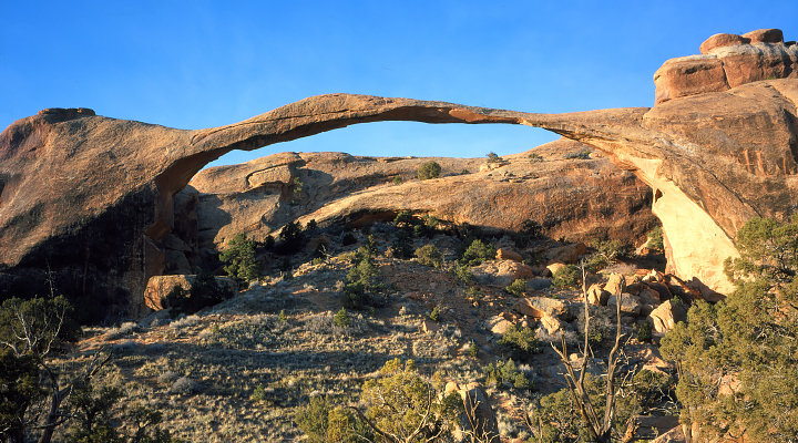 A photograph of Landscape Arch taken from the ground shows a long rock arch with blue sky, trees, and brush visible through it.