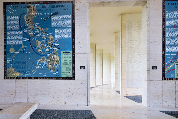Lists of names on a tablet from the Walls of the Missing are visible in the background of a photograph of the cemetery. In the foreground, a blue mosaic map depicts troop movements during a battle for the Philippines.
