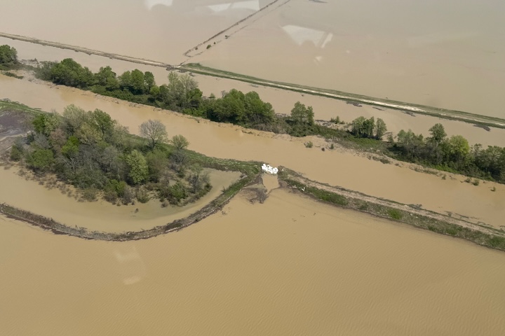 Several white sandbags fill a gap in an earthen levee in this aerial photograph. Trees line the edges of the river, and muddy floodwaters spread across the surrounding landscape.