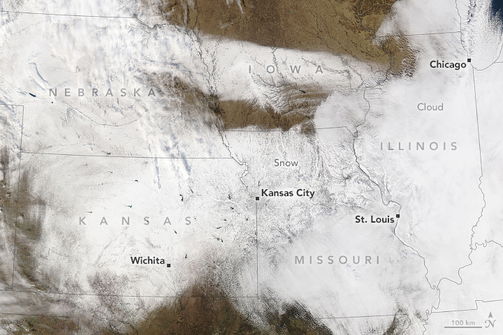 A satellite image centered on the Central U.S. is covered with a strip of white where snow fell during a storm. White clouds obscure part of the snowy scene in the left and right sides of the image.