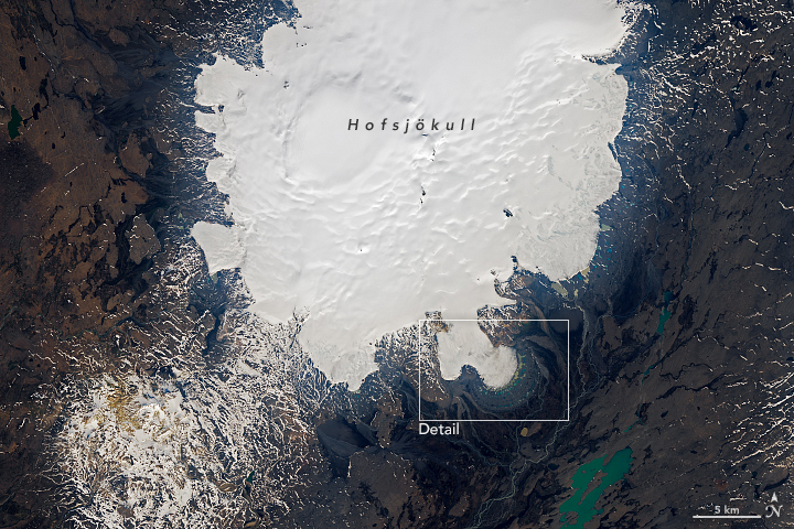 A wider view of the scene shows the glacier descending from the bottom-right side of a large, white ice cap. The ice cap spans the middle of the image and is surrounded by a brown landscape punctuated with blue-green lakes and white snow.