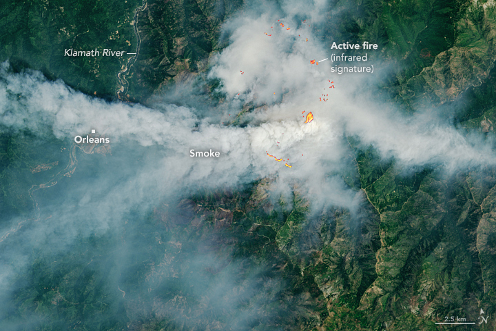 Smoke from a fire forms a distinct plume across the middle of the image and diffuse plumes toward the top-right and bottom-left. The terrain is green and mountainous, with several brown, bare areas.