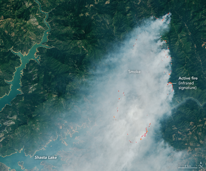 A detailed view of the first image shows the active fire area. The infrared signature of actively burning fires appears bright orange, primarily across the right half of the scene. Gray-white smoke surrounded by green forested land occupies most of the image. A few arms of a green-blue lake are visible to the left.