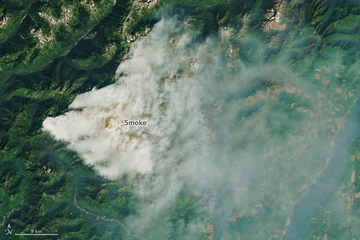 A satellite image shows mountainous terrain with a plume of smoke blowing east in a V shape. The smoke appears to concentrate in the valleys.