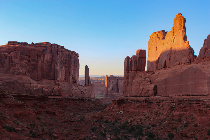 A photograph taken from the ground shows the Tower of Babel and Queen Victoria Rock standing as tall walls of red rock on the edges of a stream valley. Blue sky is visible toward the top of the photo.