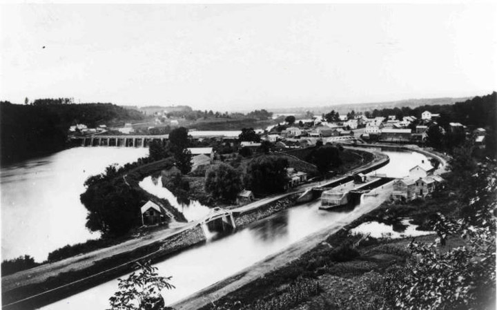 A black-and-white photograph looks down on a stretch of the Erie Canal running toward the bottom left of the frame. There is a lock in the middle of the canal, as well as a feeder canal and a wider river to the left. A stone aqueduct spans the river in the background.