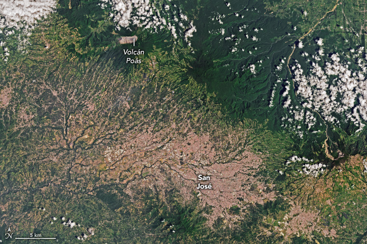 A satellite image shows the Costa Rican capital of San José and surrounding developed areas, which appear pink or tan and occupy the bottom portion of the image, and light and dark green mountainous areas across the top of the image. Volcán Poás (Poás volcano) is at the top of the frame near the center, identifiable as a mountain-top free of vegetation.