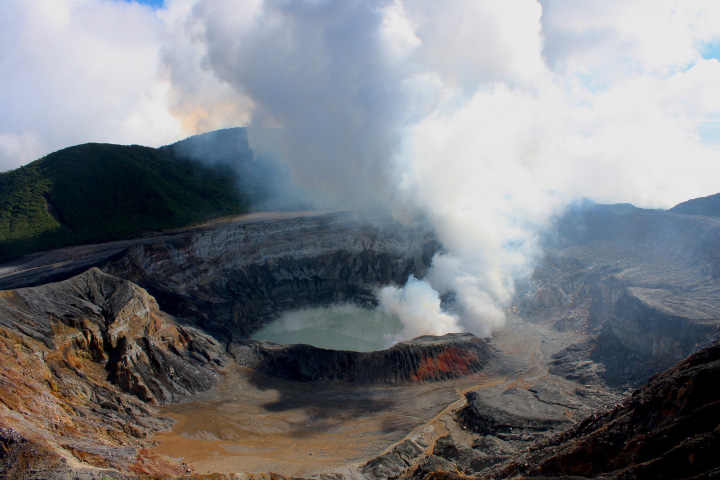This photograph was taken from the rim of the summit crater of Poás volcano. A small, light-teal lake fills in part of the crater floor, and the white billows of a gas plume rise from it.