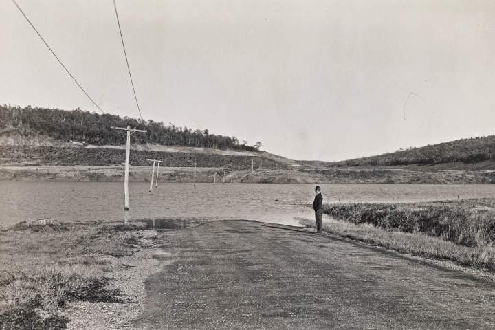 A black-and-white photograph taken in 1939 shows a person standing next to a flooded road. T-shaped electric wires protrude from the flooded area showing where the road leads.