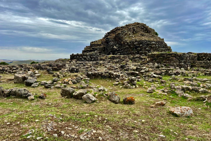A photograph taken from the ground shows the remains of a nuraghe, a domed structure made of stones, against a cloudy sky. The grassy foreground is strewn with stones of various sizes.